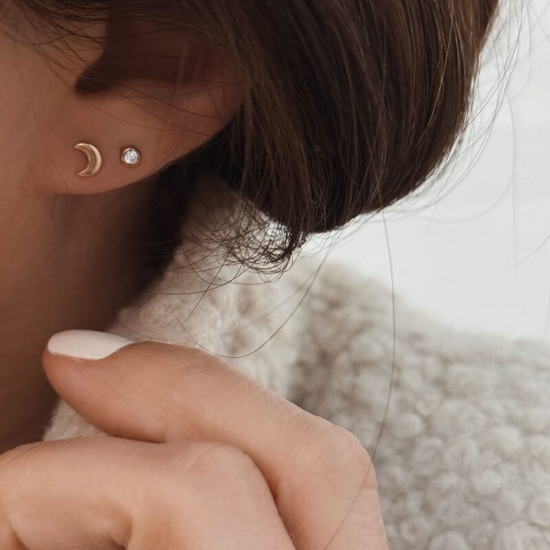 Boucles d'oreille pour femme en plaqué or au style minimaliste. Le mannequin porte une petite paire de boucles en forme de croissant de lune et une autre paire style puces rondes sertie d'un oxyde de zirconium. Une association toute en finesse et élégance.