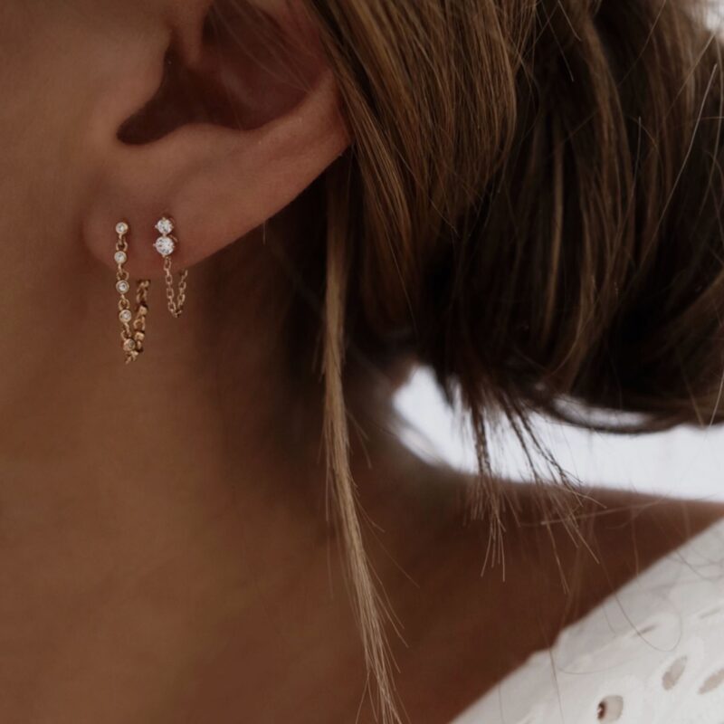 Boucles d'oreilles pour femme portées en association. Les boucles d'oreilles sont pendantes et en plaqué or 3 microns 18 carats. La première boucle est composée d'une chaîne sertie de petits oxydes de zirconium ronds. Le second bijou se compose de deux oxydes de zirconium et d'une fine chaînette qui vient pendre en dessous.