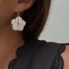 Boucles d'oreilles pour femme en acier inoxydable doré qui se composent d'une fine créole décorée d'une fleur blanche nacrée. La créole mesure 2,5 centimètre de diamètre. Fermoir crochet.