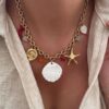 Collier pour femme en acier inoxydable doré qui résiste à l'eau. Le bijou est composé d'une chaîne à larges maillons sur laquelle sont disposés plusieurs charms : étoile de mer, coquillage en nacre, coeur en nacre, un corail rouge etc... Tour de cou ajustable.