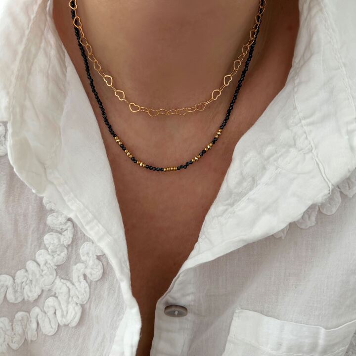 Association de deux colliers ras-de-cou Jolie Demoiselle. Le premier collier en plaqué or 3 microns 18 carats se compose d'une chaîne en forme de coeurs ajourés. Le second bijou est entièrement composé de petites perles noires et dorées.