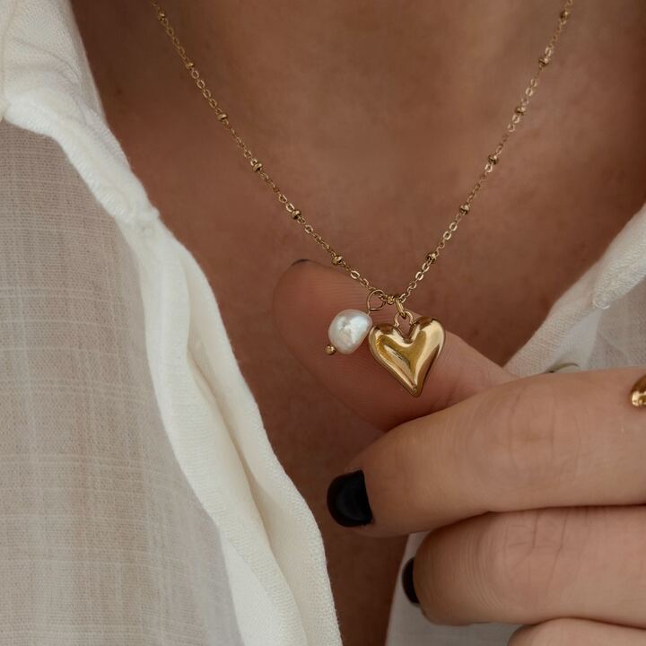Collier pour femme en acier inoxydable doré qui se compose d'une fine chaîne décoré de petites billes. La chaîne est décoré d'un petit coeur bombé et d'une perle d'eau douce. Le bijou dispose d'une chaînette de rallonge grâce à laquelle le collier est ajustable.