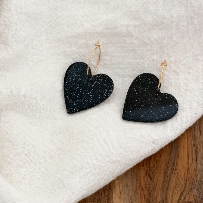 Boucles d'oreilles tendance pour femme qui se compose d'une fine créole en plaqué or 3 microns 18 carats et d'un pendentif en forme de coeur de couleur noir décoré de petites paillettes.