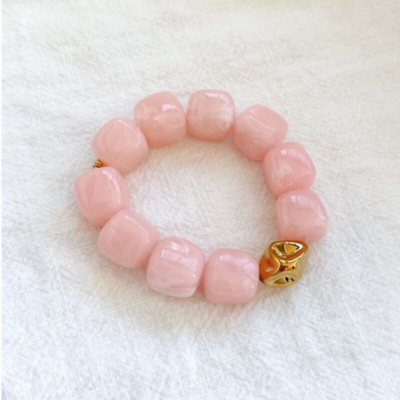 Bracelet élastique pour femme en acier inoxydable doré, très tendance. Il se compose de grosses perles roses en forme de cube et d'une perle doré en son milieu au fini irrégulier. C'est la pièce à avoir à son poignet cet été !