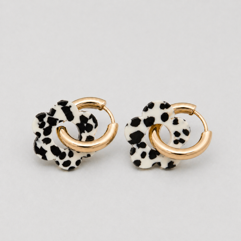 Boucles d'oreilles pour femme très tendance qui se composes d'une créole en acier inoxydable doré lisse et d'un pendentif en forme de fleur au motif Terrazzo de couleur noir et blanc. Les fleurs disposent d'un trou assez grand en leur centre afin d'être retirées de la créole et être interchangées avec d'autres fleurs ou composants.